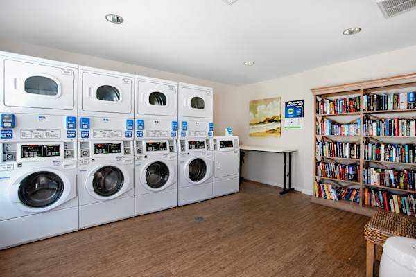 A laundry room with a row of washers and dryers, a bookshelf, and a table with a painting on the wall.