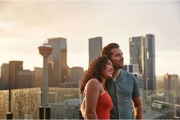 A couple is standing close together on a rooftop with a city skyline in the background.