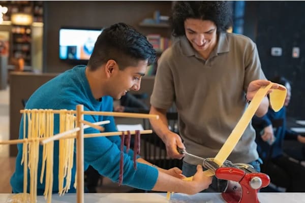 A man in a blue shirt is cutting a long piece of pasta with a pair of scissors.
