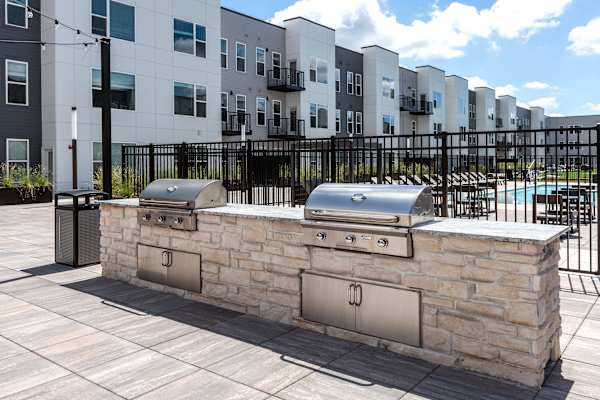 A stone barbecue grill is in front of a black fence and apartment building.