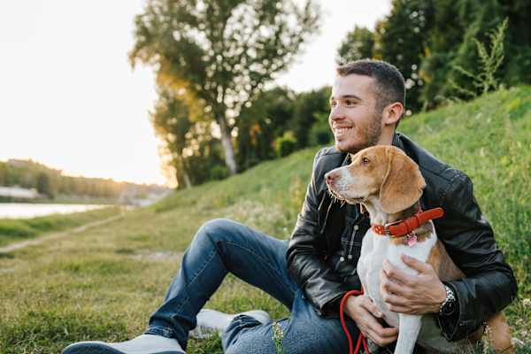 A man in a leather jacket is sitting on the grass holding a dog.