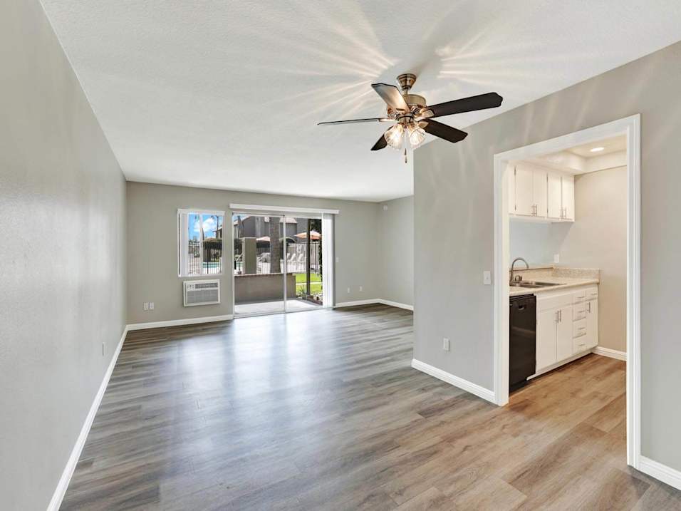 A spacious room with a ceiling fan and a kitchen area visible through the door.