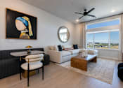 Thumbnail 2 of 18 - A model apartment living room with large windows,  furniture and a ceiling fan at Amara of a woman, a black sofa, a white couch, a coffee table, and a view of the city.