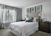 Thumbnail 3 of 33 - Bright bedroom at River Oaks at Rochester Hills Apartments with white bed linens, two nightstands, lamps, and window blinds.
