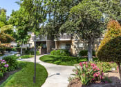 Thumbnail 23 of 25 - Grounds with lots of flowers, foliage, trees, lamp and sidewalk leading up to tan apartment building with patios/balconies.