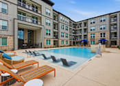 Thumbnail 9 of 55 - A large swimming pool surrounded by sun loungers and umbrellas in front of a multi-story apartment building.