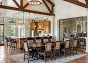Thumbnail 37 of 41 - A large wooden dining table with chairs is set up in a room with a kitchen in the background.