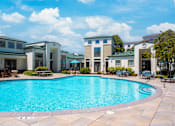 Thumbnail 3 of 31 - A swimming pool surrounded by a patio with chairs and a building in the background.