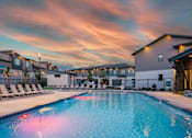 Thumbnail 2 of 26 - A swimming pool surrounded by lounge chairs and buildings under a colorful sky.