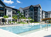 Thumbnail 1 of 59 - A swimming pool surrounded by lounge chairs and umbrellas in front of a row of apartment buildings.