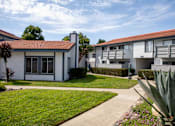 Thumbnail 19 of 22 - A white house with a red tile roof is surrounded by green grass and a concrete pathway.