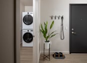 Thumbnail 11 of 34 - A white washing machine is in a small room with a black door and a plant on a table.