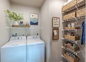 Thumbnail 5 of 26 - A small laundry room with a washer and dryer and a shelf with baskets on it.