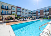 Thumbnail 1 of 30 - A swimming pool surrounded by lounge chairs and umbrellas in front of apartment buildings.