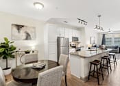 Thumbnail 2 of 29 - An interior view of a modern kitchen with a kitchen island countertop, a dining area with a dining table and chairs, and views of a living room with two large windows, a ceiling fan, and a grey couch at Parkstone at Knightdale apartments for rent in Knightdale, NC.
