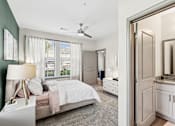 Thumbnail 6 of 29 - An interior view of a bedroom with a bed, a side table with a lamp on it, two large windows with curtains, a closet door, and a bathroom with a mirror and a granite countertop at Parkstone at Knightdale apartments for rent in Knightdale, NC.