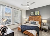 Thumbnail 5 of 29 - An interior view of a bedroom with a bed, two side tables with lamps on them, a ceiling fan, a front end table, and two large windows with curtains at Parkstone at Knightdale apartments for rent in Knightdale, NC.