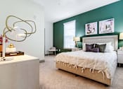 Thumbnail 7 of 29 - An interior view of a bedroom with a bed, two side tables with lamps on them, a window, and a front end table with books and a lamp at Parkstone at Knightdale apartments for rent in Knightdale, NC.
