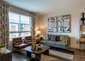 Thumbnail 4 of 26 - A living room with a grey couch, a brown chair, a coffee table, and a painting on the wall.