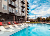 Thumbnail 13 of 15 - A large swimming pool in front of a building with red lounge chairs.