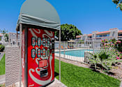Thumbnail 64 of 94 - A red vending machine with a grey roof is in the foreground of a grassy area with a pool and buildings in the background.