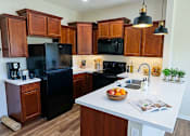 Thumbnail 2 of 29 - A kitchen with black appliances and wooden cabinets. A bowl of oranges sits on the counter beside an open magazine. Plants, Oils, a coffee maker, and various household items decorate the top of the counter.