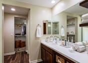 Thumbnail 7 of 29 - A bathroom with a white counter top adorned with towels, toothbrushes, and lotions next to a white sink. A walk in closet can be seen in the background, containing shirts, hangers, pants, and shoes.