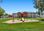 Thumbnail 16 of 17 - A playground with a swing set and sandbox in front of a building.
