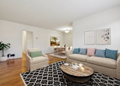 Thumbnail 10 of 23 - A living room with a beige couch, a coffee table, and a potted plant.