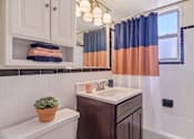 Thumbnail 7 of 24 - A bathroom with a white toilet, a brown sink, and a window with blue and orange curtains.