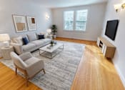 Thumbnail 4 of 17 - A living room with a grey rug, a grey sofa, a grey armchair, a wooden coffee table, a white lamp, a black TV, a plant, and two framed pictures on the wall.