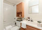 Thumbnail 11 of 20 - A white toilet sits in a bathroom with a white sink and wooden cabinets.