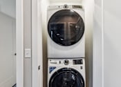Thumbnail 7 of 17 - A white front loading washing machine with a black door is in a small room.