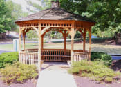 Thumbnail 20 of 21 - A wooden gazebo with a brown roof is surrounded by green bushes and trees.