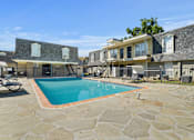 Thumbnail 6 of 10 - A large swimming pool surrounded by stone and a patio area with chairs and a table.