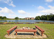 Thumbnail 8 of 14 - A picnic table sits in the middle of a grassy area with a pond and buildings in the background.