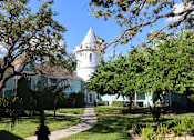 Thumbnail 21 of 23 - A white building with a tower is surrounded by green trees.