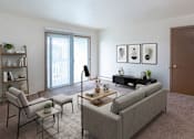 Thumbnail 2 of 11 - A living room with a grey couch, a chair, a small table with a vase and a plant on it, a bookshelf with a plant on top, and pictures on the wall.