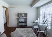 Thumbnail 9 of 14 - A living room with a grey couch, a glass table, and a television on a stand.