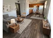 Thumbnail 4 of 4 - A living room with a grey couch, a wooden coffee table, and a kitchen area in the background.