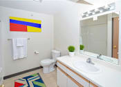 Thumbnail 5 of 14 - A bathroom with a white toilet, a white sink, a mirror, a towel rack, and a colorful rug.