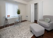 Thumbnail 9 of 17 - A room with a grey chair and ottoman, a desk with a plant on it, and a patterned rug.