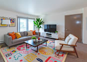 Thumbnail 9 of 13 - A living room with a grey couch, a coffee table, a chair, a plant, and a TV.