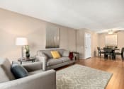 Thumbnail 8 of 41 - Apartments for Rent in Mar Vista, CA - Vista - Furnished Living Room with Grey Sofa and Armchair, Light Wood Flooring, Tan and White Carpet, Wooden Side Table with Lamp, Beige Walls, and Dining Nook with Dark Table and Chairs