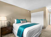 Thumbnail 15 of 41 - Mar Vista CA Apartments - Vista - Furnished Bedroom with Plush Carpeted Floors, Bed, Wooden Nightstand with Drawers and a Lamp, Beige Walls, and Large White Closet