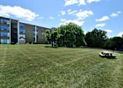 Thumbnail 6 of 12 - A large grassy field with a picnic table and apartment buildings in the background.