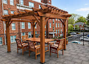Thumbnail 9 of 17 - A wooden pergola with a table and chairs is set up on a patio.
