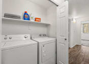 Thumbnail 8 of 25 - A laundry room with a white washer and dryer, a shelf with towels and cleaning products, and a doorway leading to another room.