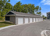 Thumbnail 29 of 53 - A two-story garage with a black roof and white doors is surrounded by a gravel driveway and a grassy area with trees.