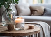 Thumbnail 2 of 11 - A candle sits on a wooden table with a cushion and a vase in the background.